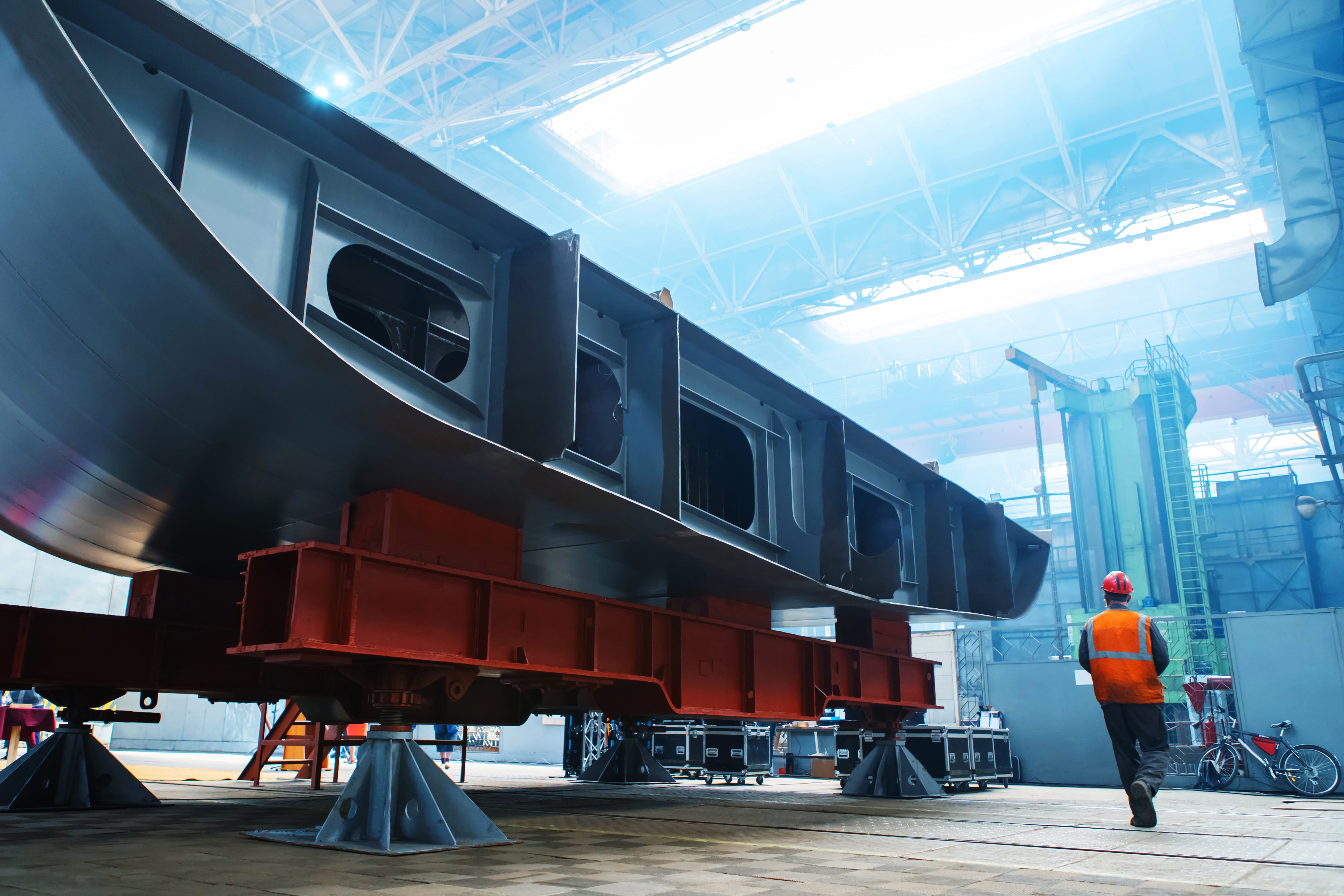 Worker in safety gear walking beside large steel frame under construction in industrial workshop