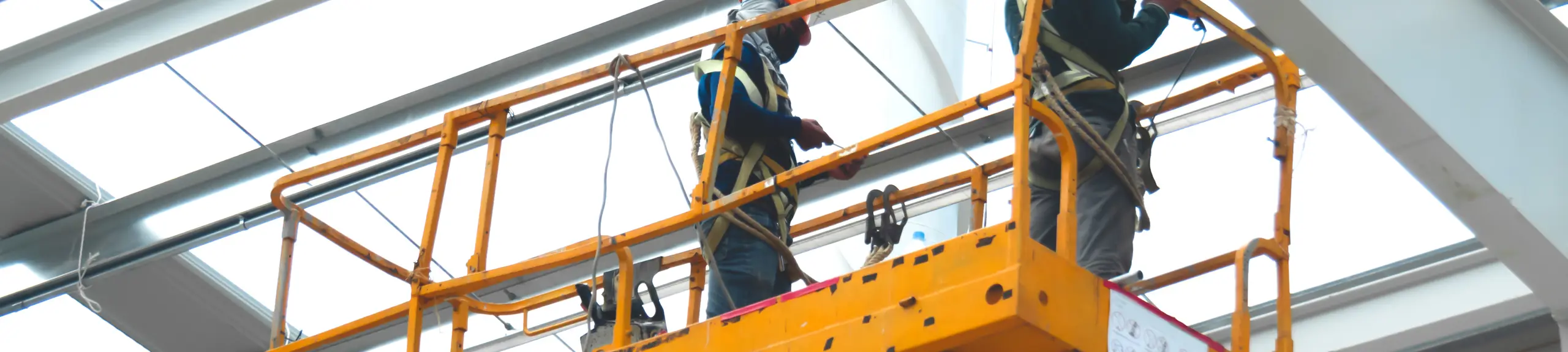 Indoors orange hydraulic lift with two workers high up wearing protective harnesses and orange hard hats. Indoors orange hydraulic lift with two workers high up wearing protective harnesses and orange hard hats.