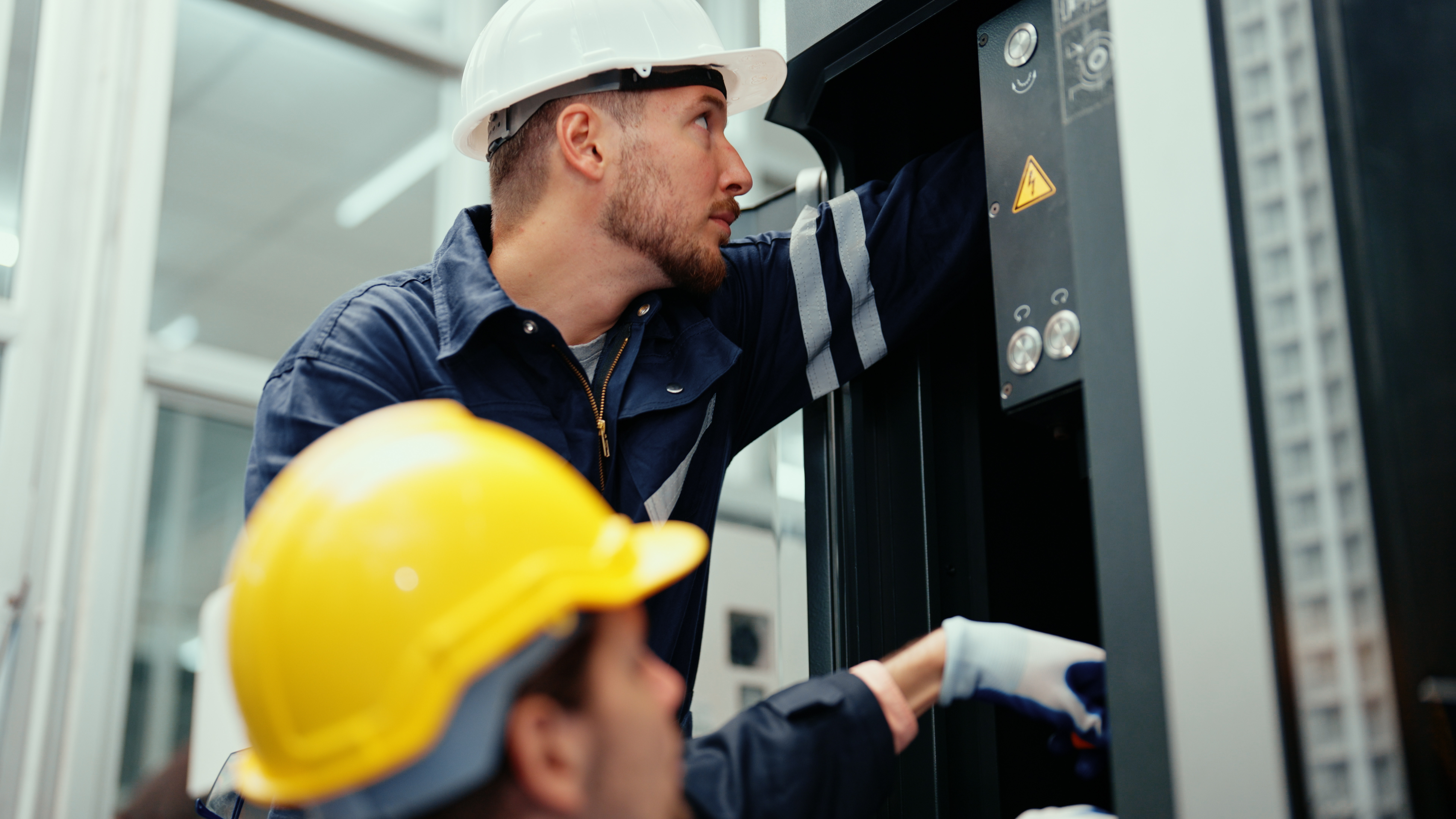 Two engineering specialists in striped blue overalls and hard hats performing safety tests on industrial equipment.